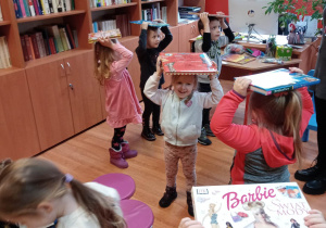 Dzieci chodzą z książkami na głowach. Uśmiechają się.