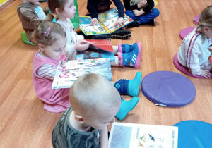 Dzieci siedzą na pufach. Trzymają na kolanach otwarte książki. Trzymają na nich palce. Czytają je.