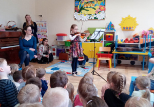 Dziewczynka stoi na środku sali, gra na skrzypcach. Przed nią siedzą dzieci i słuchają. Za nią jest dekoracja przedszkolna i szafki.
