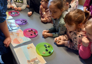 Dziewczynki i chłopcy opierają się o stolik. Na nim stoją kolorowe talerzyki z guzikami oraz obrazki bajek.