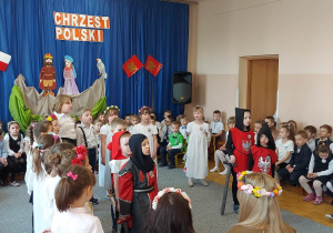 Dzieci w strojach galowych i średniowiecznych śpiewają piosenkę o Polsce.