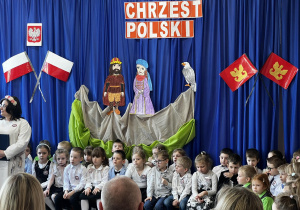 Dzieci siedzące na scenie. W tle scenografia z napisem Chrzest Polski.