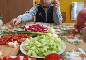 Na stole stoją talerze z pokrojonymi warzywami. Dzieci nakłuwają warzywa na patyczki od szaszłyków.