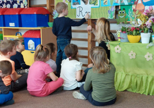 Dzieci siedzą przed tablicą. Jedno dziecko stoi przy tablicy. Kreśli palcem literę "r" po śladzie.