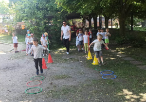 Dzieci są w ogrodzie przedszkolnym. Skaczą z obręczy do obręczy. Są podzielone na dwie drużyny.