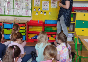 Dzieci siedzą na dywanie. Nauczyciel wskazuje na obrazki. Dzieci patrzą.
