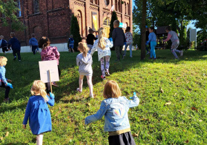 Dzieci biegają po trawie. Szukają odpadów. W tle widać kościół.