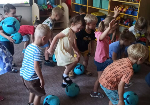 Dzieci są w sali przedszkolnej. Turlają piłkę stopami.