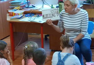 Dzieci siedzą na dywanie. Przed nimi siedzi Pani Bibliotekarka. Trzyma w ręku ilustrację. Tłumaczy dzieciom.