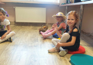 Trzy dziewczynki uczestniczki konkursu siedzą na podłodze na pufach. Uśmiechają się. Jedna macha rączką.