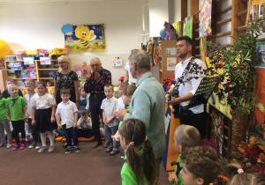 Dzieci stoją w półkolu. Między nimi stoją osoby z Senior - Vigor. Prowadzący gra na gitarze. Wszyscy śpiewają piosenkę.