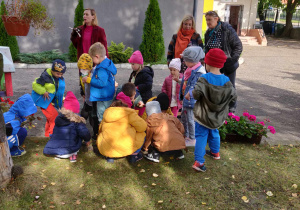 Dzieci gromadzą się w jednym miejscu. Kucają na trawie. Nauczyciele stoją za dziećmi. Obserwują dzieci