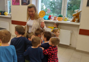 Pani nauczycielka stoi na środku. Trzyma w ręku żółtą, uśmiechniętą buźkę. Dzieci stoją przed nauczyceilką. Przytulają się do niej.