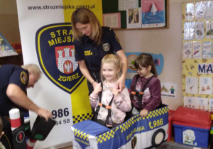 Strażniczka Miejska pomaga dziewczynkom założyć pojazd Straży Miejskiej na szelkach