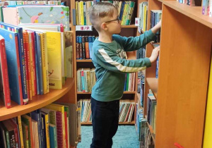 Chłopiec jest w bibliotece. Patrzy na półkę z książkami. Wyjmuje książkę z półki.