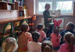 Nauczycielka siedzi na krzesełku. Trzyma w ręku książkę. Dzieci siedzą na podłodze. Patrzą na nauczycielkę.