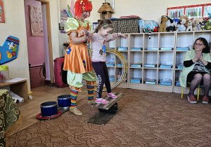 Dziecko z pomocą cyrkowca stoi na równoważni.