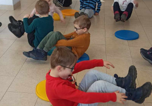 Dzieci siedzą na kolorowych pufach. Mają podniesione ręce i nogi.