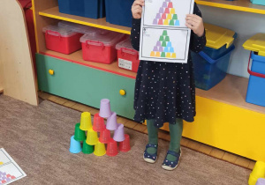Dziewczynka stoi na dywanie w klasie. Trzyma w rękach kartkę z wydrukowanymi kolorowymi kubkami. Obok niej stoi budowla z kubeczków.