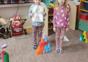 Dwie dziewczynki stoją na dywanie. Jedna znich trzyma kartkę z wydrukowanymi kolorowymi karteczkami.Między nimi stoi nudowla z kolorowych kubeczków.
