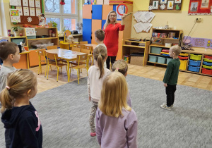 Dzieci stoją w rozsypce przed nauczycielką. Nauczycielka stoi przed dziećmi. W ręce trzyma tamburyno.
