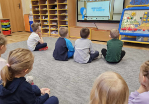 Dzieci siedzą na dywanie przed tablicą multimedialną. Oglądają film edukacyjny Niecio sam w domu.