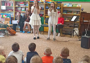 Dwie dziewczynki stoją na środku sali. Trzymają mikrofony w rękach. Śpiewają piosenkę. Siedzą przed nimi przedszkolaki, widownia.