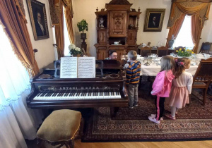 Na zdjęciu jest otwarty fortepian. Widoczne jest również starodawne wnętrze mieszkalne. Stoją tam dzieci.