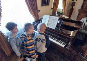Trzech chłopców stoi przy fortepianie. Ogląda go.