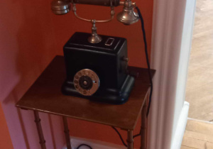 Na zdjęciu jest starodawny telefon z słuchawką na widełkach. Kolor czarny.