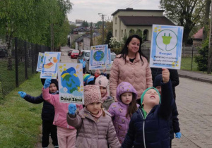 Dzieci idą chodnikiem. Są ubrane w kurtki i czapki. W rękach trzymają transparenty z hasłami ekologicznymi.