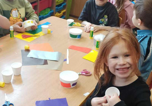 Dzieci siedzą przy stoliku. Uśmiechają się. Wycinają elementy z kolorowego papieru. Przyklejają do pojemniczków.