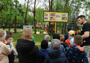 Dzieci stoją przed tablicą informacyjną o pszczołach murarakach. Nauczyciel opowiada o pszczołach.