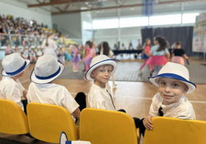 Chłopcy siedzą na krzesełkach żółtych. Mają na głowach kapelusze. Dwóch z nich odwraca się. Uśmiechają się.